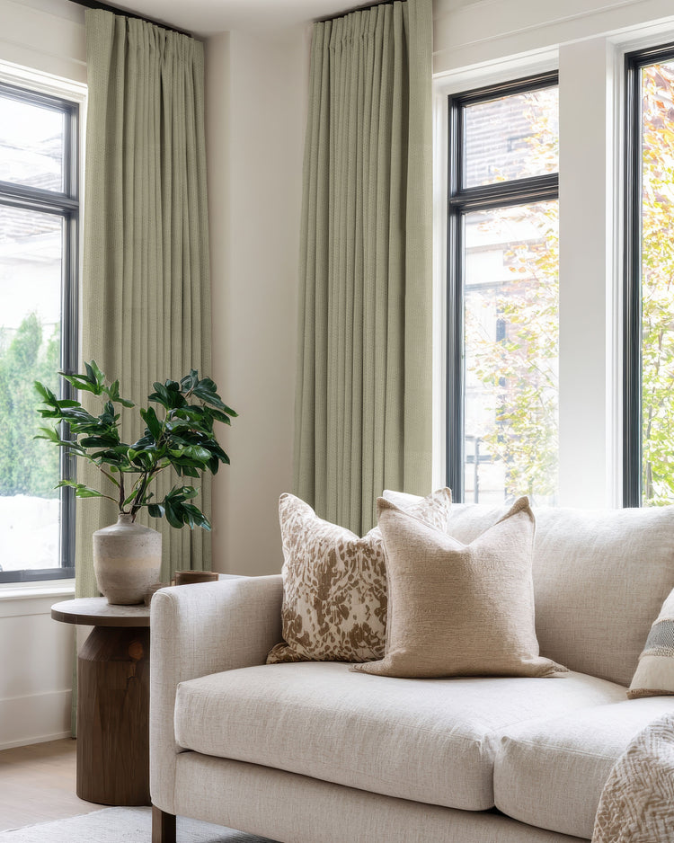 Sage blackout linen-look custom pleated curtains flank large windows in a sophisticated living room, offering privacy & light control.